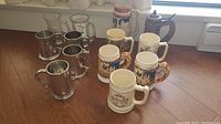 Photo showing the full lot of nine beer steins and mugs arranged on wooden floor including ceramic steins, metal tankards, and glass mugs