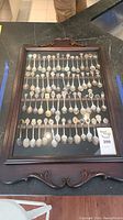 Framed wooden display case with decorative carvings containing about 56 collector spoons with various designs inside, photographed from front with glass closed.