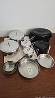 Full view of the cookware set including stainless steel pots with lids, pans, black speckled enamel pots, and wooden cutting board underneath