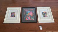 Three framed Dean Russo abstract dog art prints displayed lying flat on a wood floor. Two have white frames, one black frame.