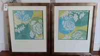 Two framed prints placed side by side showing floral designs in blue, green, and yellow with white flowers and leaves, in light wood frames with wide white mats.