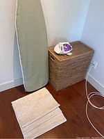 Photo showing Ridgfid ironing board leaning against the wall with a small purple and white iron placed on a woven hamper next to beige rugs on the floor.