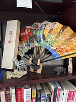 Japanese folding fan displayed open with multi-color floral and cartwheel design, shown with small statues arranged beside it and the fan's storage box behind.