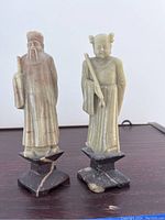 Front view of two small carved stone monk statues on black stone pedestal bases showing distinct features and coloration of each.