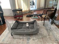 Pet bed inside a metal pen, positioned on area rug with sunlit windows in background.