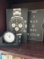 Rolex hardcover book and black desk clock displayed on a shelf.