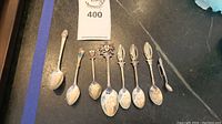 Seven sterling silver souvenir spoons arranged in a row showing detailed and varied decorative handles on a black surface.