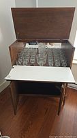 Vintage wooden bar cabinet with open lid showing assortment of clear etched glassware inside and pull-out surface panel below.
