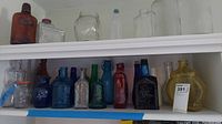 Shelf with multiple figural glass bottles in assorted colors including clear, amber, blue, green, red, and yellow. Bottles vary in shape and size, some with embossed decorations.