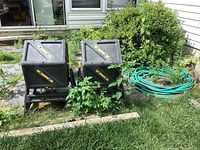 Photo of two black Miracle-Gro composters on stands with yellow handles, showing weathering and vegetation around them, with green garden hose coiled nearby outside a house.