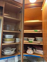 Open kitchen cabinet showing Corningware casseroles, bowls, and teapot inside shelves