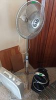Photo showing the metal floor fan, black dual fan heater, and white box fan grouped together on carpeted floor near wood wall.