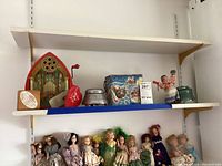 Shelf displaying vintage music boxes including red tin crank music box with church design, heart-shaped music box, metal Victorian scene boxes, and a rectangular winter scene tin. Below shelf, a group of vintage dolls.