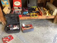 Photo showing overall assortment of power tools and hand tools arranged on a wooden shelf and concrete floor, including cases and boxed items.