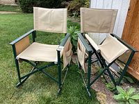 Two outdoor folding rocker chairs on grass, beige canvas with green metal frames, showing age and dirt.