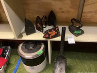 Overview photo showing all five sad irons including the painted one, the stoneware crock with lid, and the fireplace ash shovel
