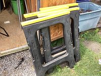 Four black plastic folding saw horses stacked and folded, showing yellow top edges and textured sides.