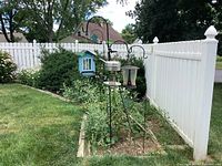 Four black metal shepherd hooks with assorted bird feeders hanging outdoors by garden bed and white fence.