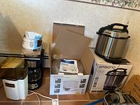 Four small kitchen appliances on kitchen floor against wall: Ambiano electric pressure cooker on box, Ambiano food dehydrator in open box, Presto popcorn maker, and Hamilton Beach coffee maker with coffee filters on top.
