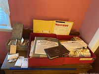 Wide view of a table with a Hallmark box full of used stamps and envelopes, a large red box container with loose stamps and envelopes, and a bicentennial commemorative coin album placed on top.