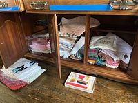 Wooden cabinet open showing multiple folded tablecloths, placemats, and linens with different colors, patterns, and textures stacked inside and some on the floor.
