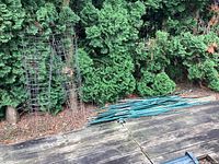 Wide view of 11 metal tomato cages stacked against green shrubbery, and a separate pile of assorted green metal garden stakes and fencing posts laying on wood deck