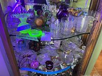 Cabinet view under UV light showing various glass items including green glowing uranium glass vase, clear pressed glass tumblers, purple hobnail vase and dried floral arrangement.