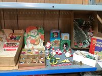 Overview of assorted Christmas ornaments, figurines, and decorations on shelf including boxed and loose ornaments, paper decorations and small figurines
