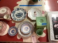 Overview of pink rose patterned dishes, advertising plates, green glass canister with lid and Fire King cup seen on red and beige table surface