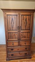 Front closed view of tall wooden armoire with 5 drawers and decorative panel doors