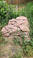 Photo of stacked flag stones outdoors on grass and dirt, near fence and plants.