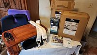 Photo showing two boxed king comforter sets, twin sheet sets, wooden bed tray, seat cushions, heating pads, candles, and masks arranged on a surface.