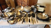 Full view of all pots, pans, pressure cooker, slow cooker, cookware strainers, grater, lids, and cooking utensils arranged on a table