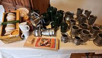 Full view of all items in the lot on a table, showing green canisters, metal grater, boxed soup set, ceramic mugs, and pewter cups.