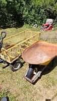 Wide outdoor photo showing yellow metal mesh wagon with black curved handle and white-walled tires next to a rusted metal wheelbarrow with wooden handles on grass