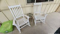 Two white wooden porch rocking chairs set side by side with small metal side table between them on a porch.