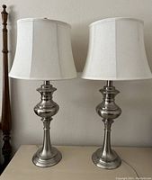 Pair of vintage Quoizel brushed nickel table lamps side by side on a table with their white fabric shades visible.