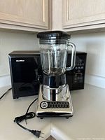 Photo showing a Cuisinart PowerBlend 600 blender with glass jar and black lid positioned in front of a black Quasar microwave.