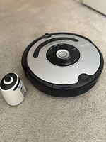 Silver and black iRobot Roomba robot vacuum cleaner on carpet, with virtual wall device beside it.