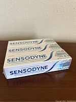 Three stacked sealed boxes of Sensodyne toothpaste in white and blue packaging, showing brand name and product details.