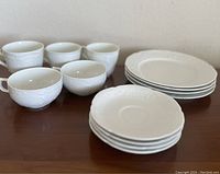 Five ivory teacups with ornate handles and embossed details, five matching saucers, and five matching small plates arranged on wooden surface.