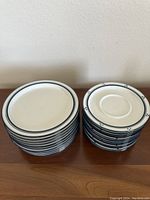 Two stacks of Dansk ceramic plates on a wooden surface, larger plates on left, smaller plates on right, showing the blue double line and dot pattern on white ceramic.