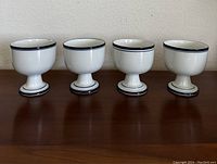 Four white ceramic goblets with blue trim, shown in a row on a wooden surface against a light wall.