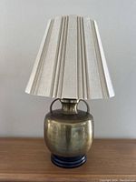 Front view of vintage brass Frederick Cooper table lamp showing pleated off-white fabric shade and rounded brass body with dual handles on black base.
