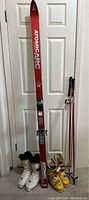 Full view of red Atomic skis standing against door, together with two pairs of ski boots and ski poles. Lot display showing all items included.