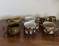 Full view of seven vintage stoneware mugs displayed on wooden surface showing various floral and abstract patterns and two handles on one mug.