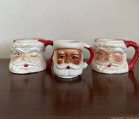 Front view of three vintage ceramic Santa mugs: two with white bearded winking faces and one with darker skin tone Santa face with eyes open. Red hat rim handles visible.