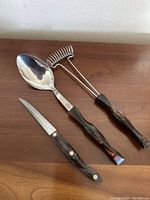 Three kitchen utensils laid out - large spoon, serrated knife, and potato masher with matching marbled handles.