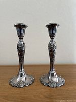 Pair of silver baroque style candlestick holders displayed upright on a wood surface against a plain background.