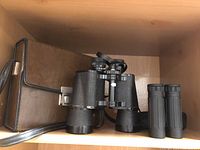 Vintage large black binoculars with textured grips and case on a wooden shelf
